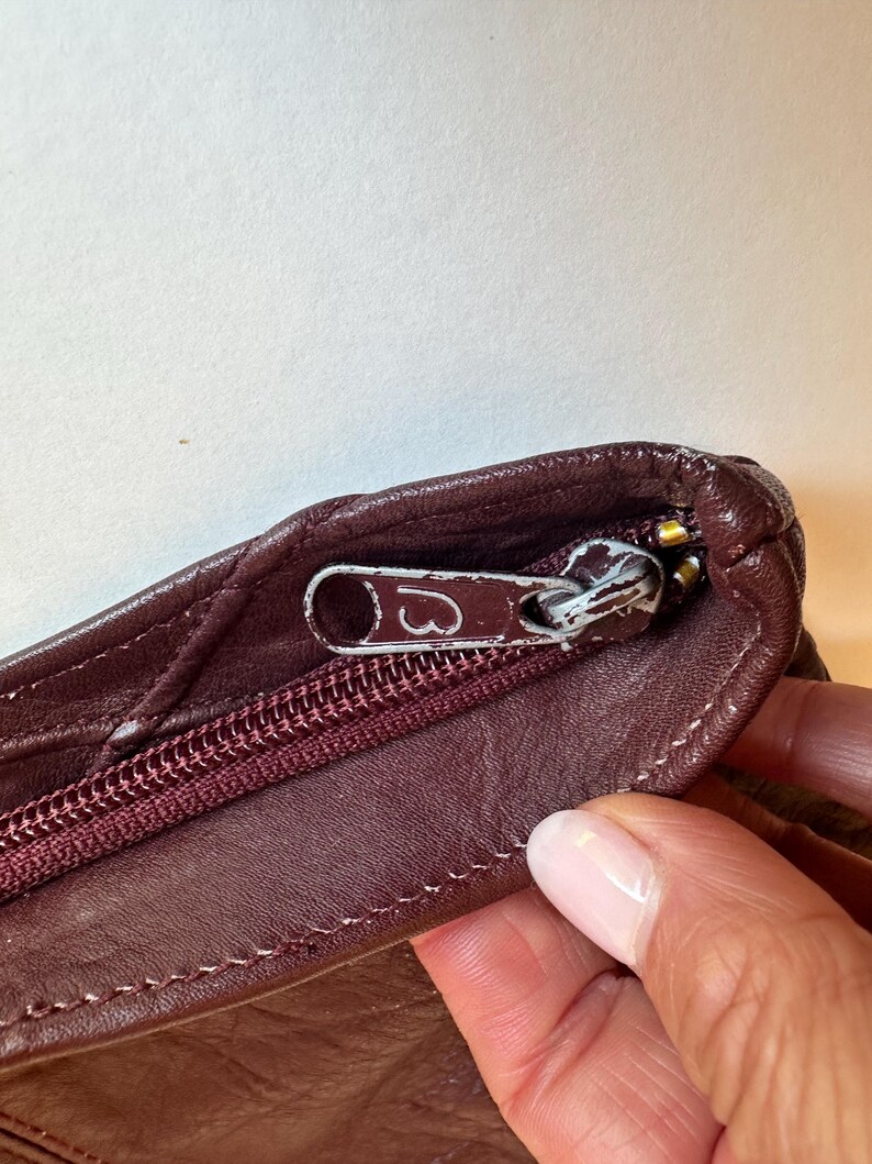 May include: Close-up of a burgundy leather bag with a silver zipper. The zipper pull has a heart design. The bag's leather has a quilted pattern and is stitched with burgundy thread. The bag is partially open, revealing the interior.