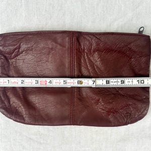 May include: A burgundy leather clutch with a zipper closure, measuring approximately 10 inches in length. The clutch has a textured surface and a simple, rectangular design. The zipper is silver.