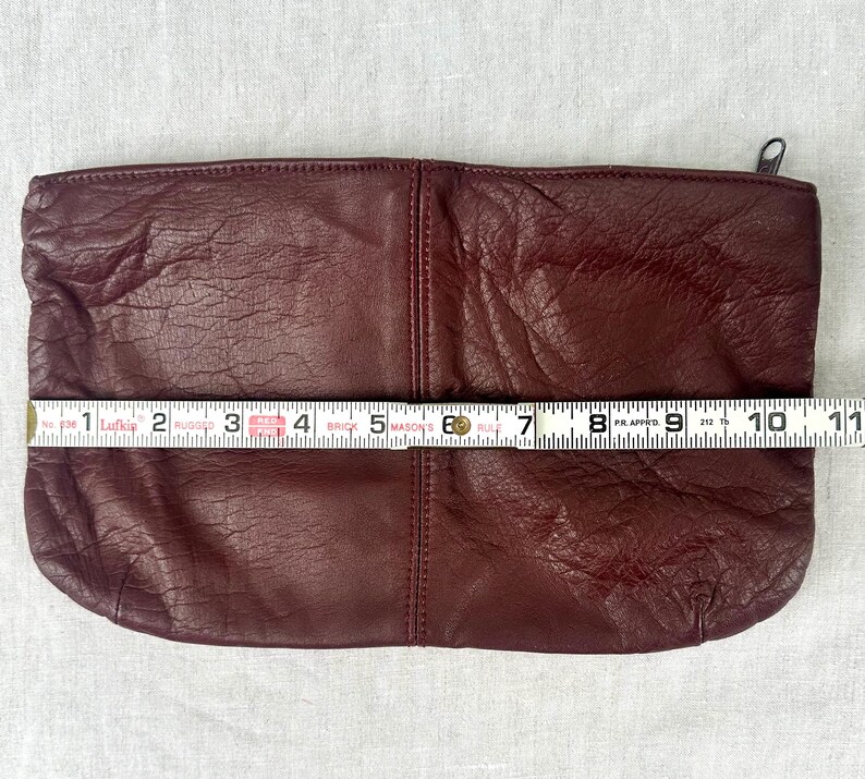 May include: A burgundy leather clutch with a zipper closure, measuring approximately 10 inches in length. The clutch has a textured surface and a simple, rectangular design. The zipper is silver.