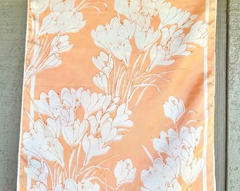 May include: Peach-colored scarf with a white floral pattern. The scarf features a repeating design of stylized white flowers and stems against a peach background. A white border frames the floral design.