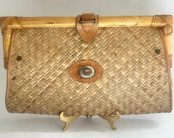 May include: A vintage woven straw clutch with a bamboo frame and leather accents. The rectangular bag features a woven pattern, a leather clasp, and a decorative oval leather patch with a silver emblem. The corners are reinforced with bamboo.