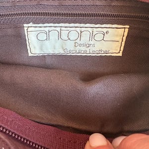 May include: Close-up of the interior of a burgundy leather bag. The bag's lining is a muted brown color. A label inside reads "antonia Designs Genuine Leather". A burgundy zipper and stitching are visible.
