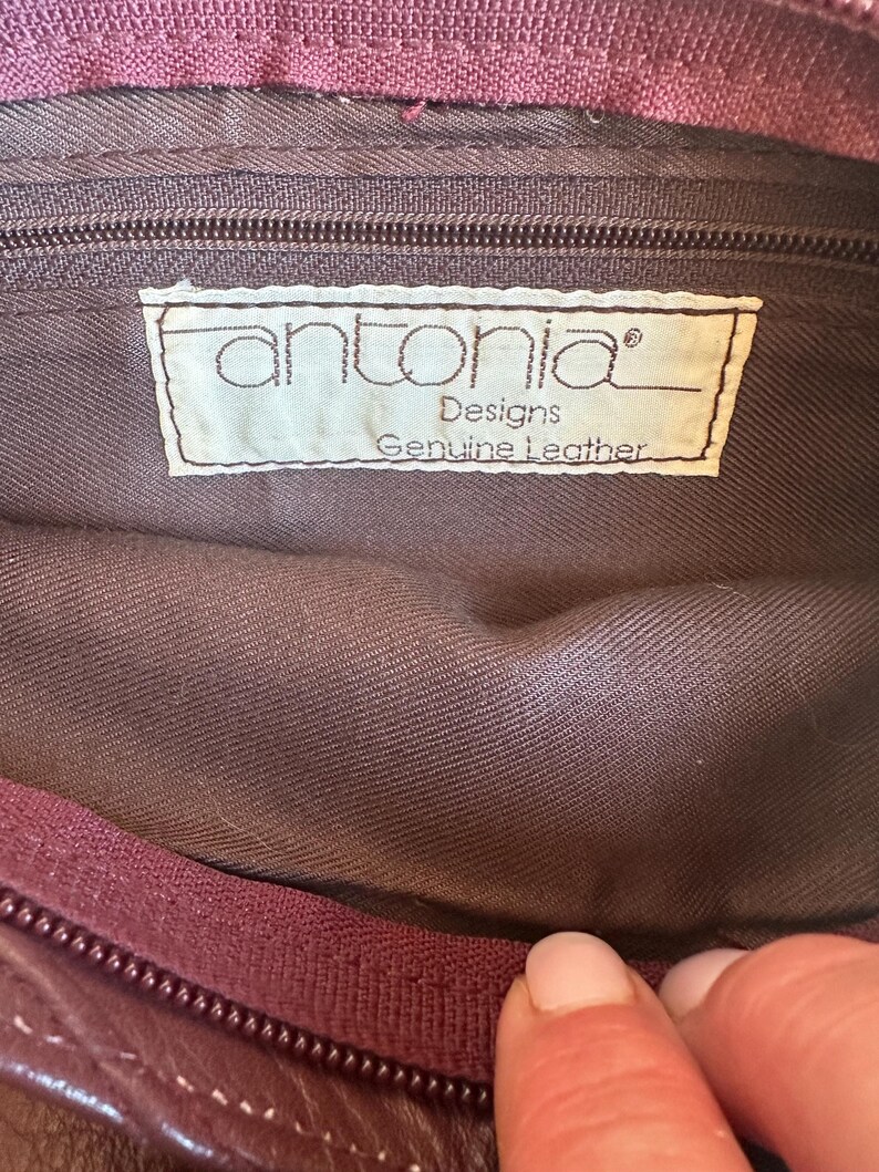 May include: Close-up of the interior of a burgundy leather bag. The bag's lining is a muted brown color. A label inside reads "antonia Designs Genuine Leather". A burgundy zipper and stitching are visible.