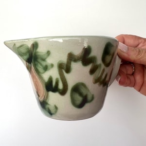 May include: A ceramic pitcher with a spout and handle. The pitcher is painted with green and brown abstract designs on a light green background. The pitcher is held by a person.