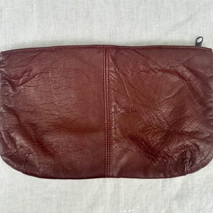 May include: A burgundy leather clutch with a zipper closure. The rectangular bag has a textured surface and a vertical seam down the center. The zipper pull is a small, dark metal.