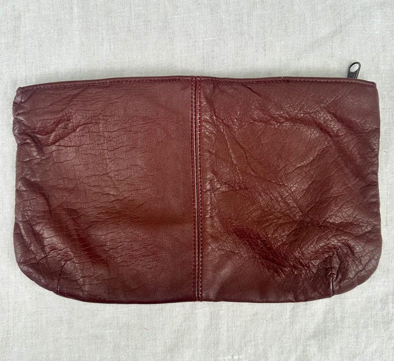 May include: A burgundy leather clutch with a zipper closure. The rectangular bag has a textured surface and a vertical seam down the center. The zipper pull is a small, dark metal.