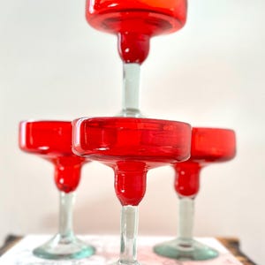 May include: A stack of four red margarita glasses with clear stems and bases. The glasses have a classic, rounded bowl shape and are made of glass. The glasses are arranged on a white and gray surface.
