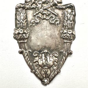 May include: A vintage sterling silver shield-shaped emblem. The emblem features ornate floral and ribbon designs, with a blank central area. The word "STERLING" is visible on the right side. The emblem has a tarnished, antique appearance.