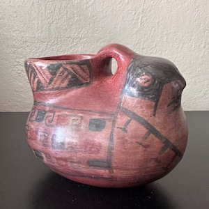 May include: A terracotta-colored ceramic vessel shaped like a bird with a handle on its back. The piece features hand-painted black geometric patterns and linear designs across its rounded body, with a distinct beak-like protrusion at the front.