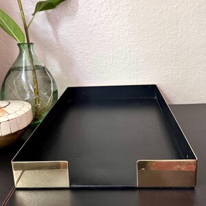 May include: A rectangular black metal tray with gold-colored accents on the short sides. The tray is empty and sits on a dark surface. A glass vase with a plant is visible in the background.