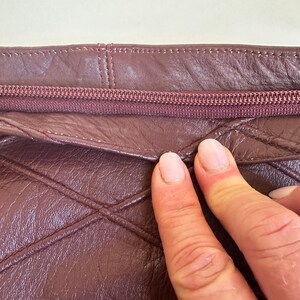 May include: Close-up of a burgundy leather bag with a quilted diamond pattern. The zipper is a matching burgundy color. The stitching is visible along the seams. The bag appears to be made of a soft, supple leather.