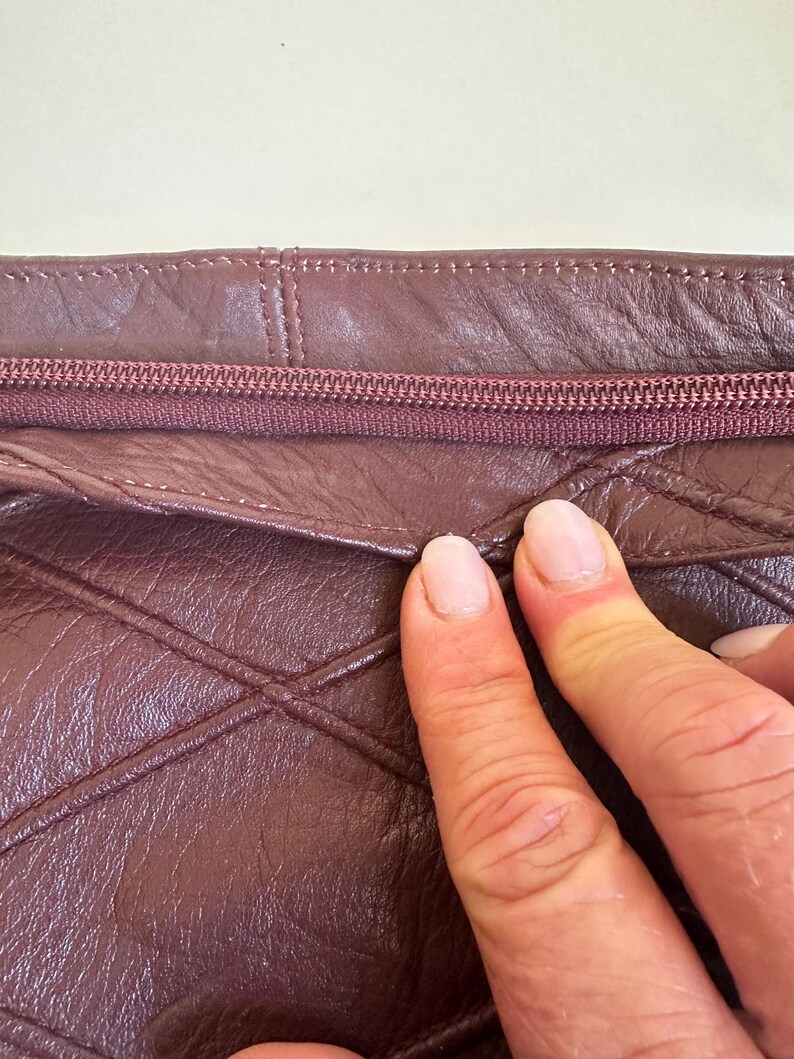 May include: Close-up of a burgundy leather bag with a quilted diamond pattern. The zipper is a matching burgundy color. The stitching is visible along the seams. The bag appears to be made of a soft, supple leather.