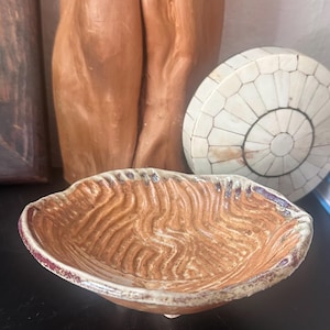 May include: A shallow, hand-formed ceramic bowl with an irregular, wavy rim. The interior features a textured, rippled surface in a warm terracotta brown, while the outer edge is finished with a speckled, cream-colored glaze and dark accents.