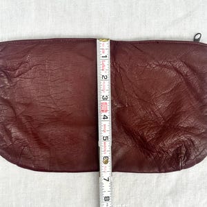 May include: A burgundy leather clutch with a zipper closure. The clutch is approximately 7 inches long. The leather has a textured appearance. A measuring tape is placed next to the clutch.