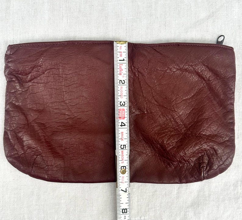 May include: A burgundy leather clutch with a zipper closure. The clutch is approximately 7 inches long. The leather has a textured appearance. A measuring tape is placed next to the clutch.