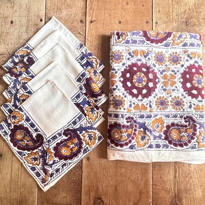 May include: A set of five square napkins and one rectangular tablecloth featuring an off-white base with a printed pattern of maroon, purple, and mustard yellow floral and paisley motifs arranged in a repeating border and central design.