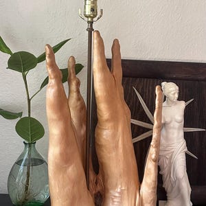 May include: A table lamp featuring a base crafted from a natural light-toned wood with a smooth, organic, multi-pronged vertical structure. A thin, dark metal rod extends upward from the center of the wood base to support a single light bulb.