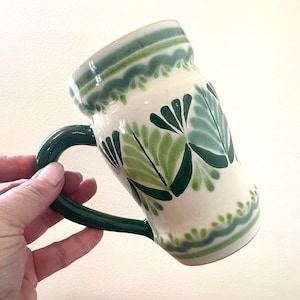 May include: A ceramic mug with a white base, adorned with green leaf patterns and decorative green bands. The mug has a dark green handle and rim, and is approximately 5 inches tall.