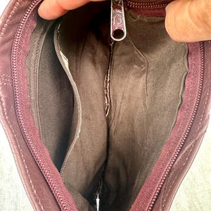 May include: Open view of a burgundy leather handbag, revealing the interior compartments. The bag features a zipper closure and a small interior pocket. The lining is a darker shade of brown. The zipper pull has a decorative design.