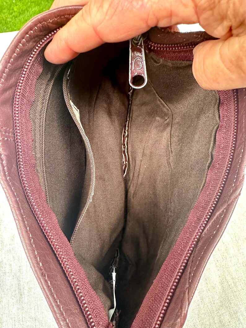 May include: Open view of a burgundy leather handbag, revealing the interior compartments. The bag features a zipper closure and a small interior pocket. The lining is a darker shade of brown. The zipper pull has a decorative design.