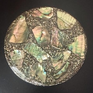 May include: A circular coaster featuring a mosaic design of iridescent abalone shell fragments embedded in a dark, textured granular base. The shell pieces display shifting green, pink, and purple tones, creating a reflective surface pattern.