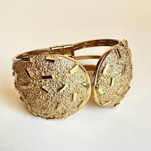 May include: A gold-toned hinged cuff bracelet. The bracelet features two large, textured, circular panels with a speckled surface and rectangular accents. The bracelet is open, revealing the hinge and inner band.