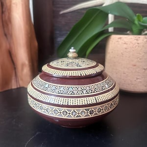 May include: A round, dark brown lidded box featuring intricate geometric inlay patterns in cream and iridescent shell. The lid is topped with a small, textured gold-toned knob and a tiny white bead. The surface has a smooth, polished finish.