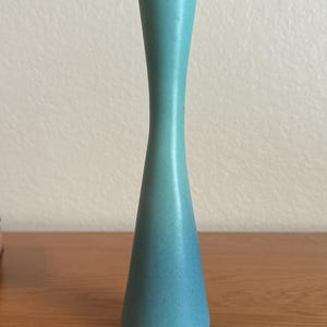 May include: A tall, slender, turquoise-colored candlestick holder with a smooth, matte finish. The object features a flared base that tapers inward toward the middle before widening slightly at the top, creating a continuous, curved silhouette.