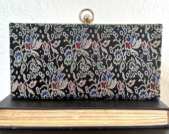 May include: A black clutch purse with a floral pattern and a gold metal clasp. The purse is sitting on top of two books.