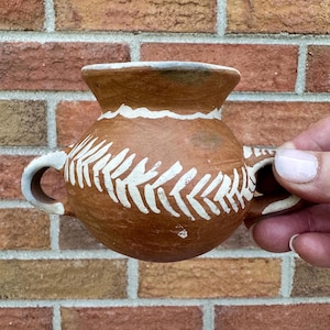 May include: A small, brown ceramic vase with two handles and a wide opening. The vase is decorated with white leaf-like patterns around the body and a white band around the rim. The vase is held against a brick background.