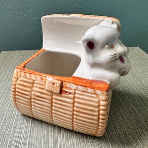 May include: A ceramic planter shaped like a woven basket with a white cat head emerging from the top. The basket is tan with orange trim and a small latch. The cat has pink accents on its ears and mouth.