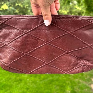 May include: A burgundy leather clutch with a diamond pattern. The clutch is rectangular and has a zipper closure. The leather appears to be soft and supple, with a slight sheen. The stitching is visible and adds to the design.