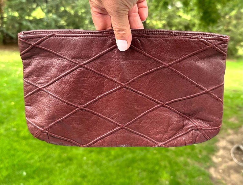 May include: A burgundy leather clutch with a diamond pattern. The clutch is rectangular and has a zipper closure. The leather appears to be soft and supple, with a slight sheen. The stitching is visible and adds to the design.