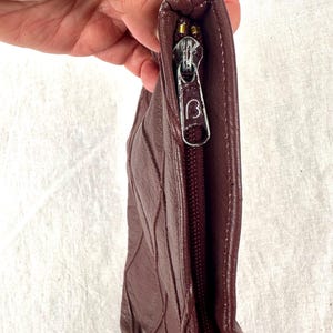 May include: A burgundy leather clutch with a quilted design. The zipper is gold-toned with a 'B' logo on the pull. The clutch is held by a hand, showcasing the texture and color of the leather.