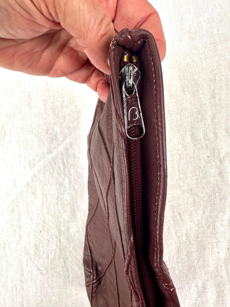 May include: A burgundy leather clutch with a quilted design. The zipper is gold-toned with a 'B' logo on the pull. The clutch is held by a hand, showcasing the texture and color of the leather.