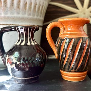 May include: Two small ceramic pitchers with handles. The left pitcher is dark brown with white vertical lines and a floral motif. The right pitcher is orange with dark brown diagonal stripes and a smooth, glossy finish on both items.
