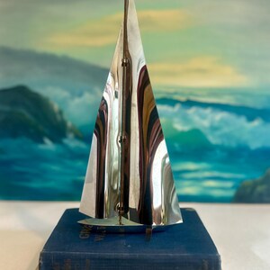 May include: A polished silver sailboat sculpture with a triangular sail, resting on a stack of two vintage books. The sail reflects the surrounding environment. The books are blue and tan, with the title "Sea Stories" visible.