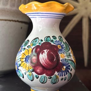 May include: A small, hand-painted ceramic vase with a white base, adorned with a large burgundy flower and colorful floral accents. The vase has a yellow rim and base, with blue stripes near the top. A decorative piece for the home.