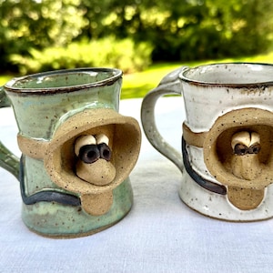 May include: Two ceramic mugs with unique monkey-like face designs. One mug is light green, the other is white. Both have a tan-colored face with black and white eyes, and a brown band around the mug.
