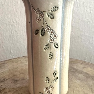 May include: A tall, off-white ceramic vase with a floral design. The vase has a rectangular shape with a slightly flared top and a light blue trim. The floral design features small flowers and leaves.