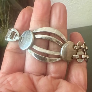 May include: A silver-toned metal bracelet featuring a central cage-like design composed of three curved bars connecting two circular discs. The bracelet includes a linked chain section on one side and a rectangular clasp mechanism on the other end.