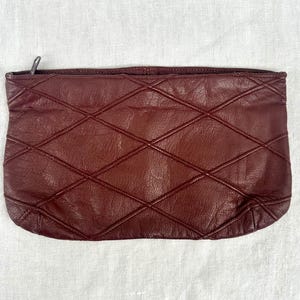 May include: A burgundy leather clutch with a diamond-stitched pattern. The rectangular bag has a zipper closure and a soft, textured appearance. The clutch is a deep, rich shade of red-brown.