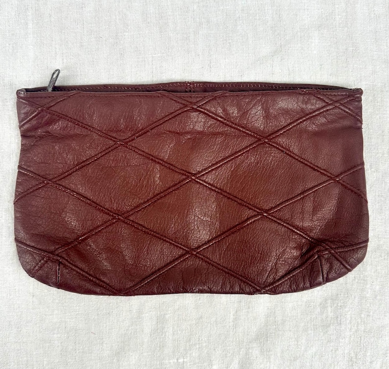 May include: A burgundy leather clutch with a diamond-stitched pattern. The rectangular bag has a zipper closure and a soft, textured appearance. The clutch is a deep, rich shade of red-brown.