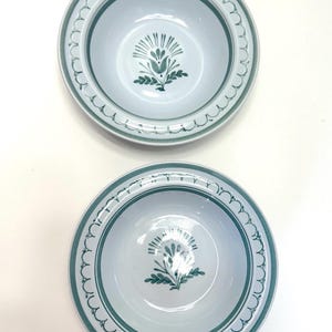 May include: Two light blue ceramic bowls with a green floral design in the center. The bowls have a green scalloped border and a green band around the rim. The bowls are likely for serving food.