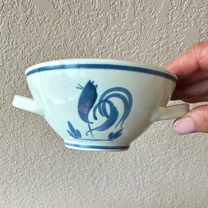 May include: A white ceramic bowl with a blue rooster design. The bowl has two small handles and a blue stripe around the rim. The rooster is depicted in a stylized, folk art style.