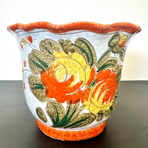 May include: A white ceramic planter with a scalloped rim and a vibrant floral design. The flowers are orange and yellow with green leaves, set against a white background. The rim and base are trimmed in orange.