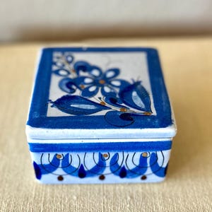 May include: A rectangular ceramic trinket box with a lid, featuring a blue and white floral design. The box has a blue border and a repeating pattern around the base. The floral design includes blue flowers and leaves with gold accents.