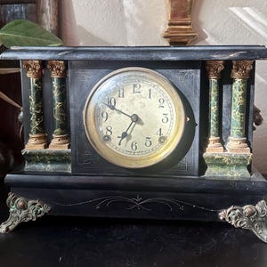 May include: A rectangular black mantel clock featuring four green marble-patterned columns with gold-toned capitals. The clock face is circular with black Arabic numerals, gold-toned hands, and ornate metal feet with a weathered finish.