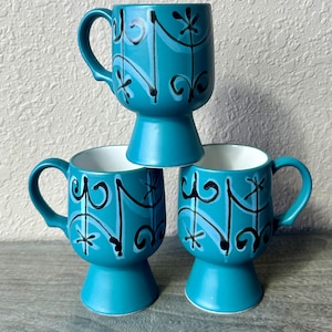 May include: Three teal blue ceramic mugs with black and white abstract designs. Each mug has a pedestal base.