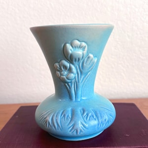May include: A light blue ceramic vase with a flared rim and a rounded base. The exterior features a raised relief design of three blooming flowers with stems and leaves extending downward toward the base of the vessel, finished in a matte glaze.